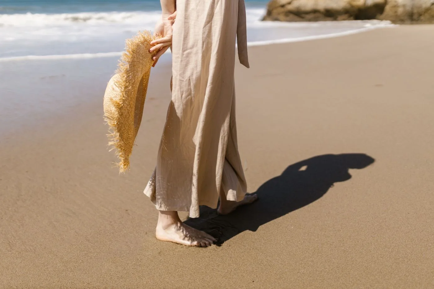 A person in a light flowing outfit carrying a straw hat on a beach