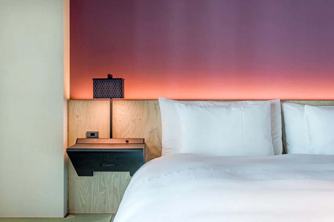 Hotel bed with white pillows beside a wooden bedside panel and warm wall lighting